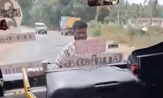 அரசு பேருந்து முன்புறம் தொங்கியபடி பயணி ஒருவர் செல்லும் காட்சி